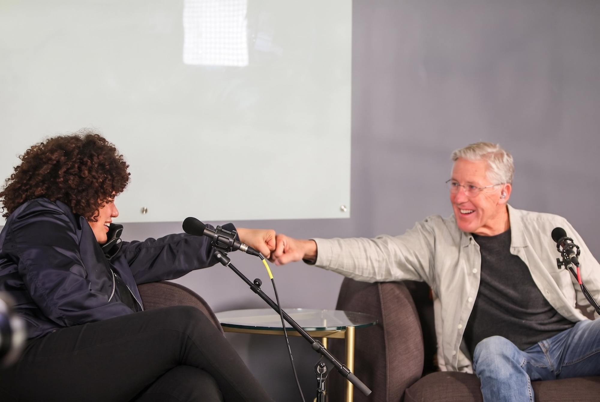Audrey Cavenecia and Pete Carroll bumping fists across the microphones.